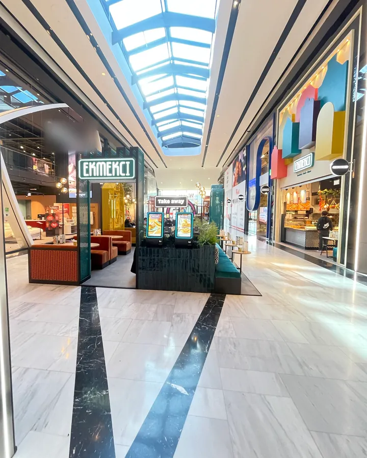 De gang van een overdekt winkelcentrum met daglicht door het glazen dak. Links de kiosk van Ekmekci met Take away en bestelzuilen, rechts de vestiging met gekleurde blokken boven het logo.