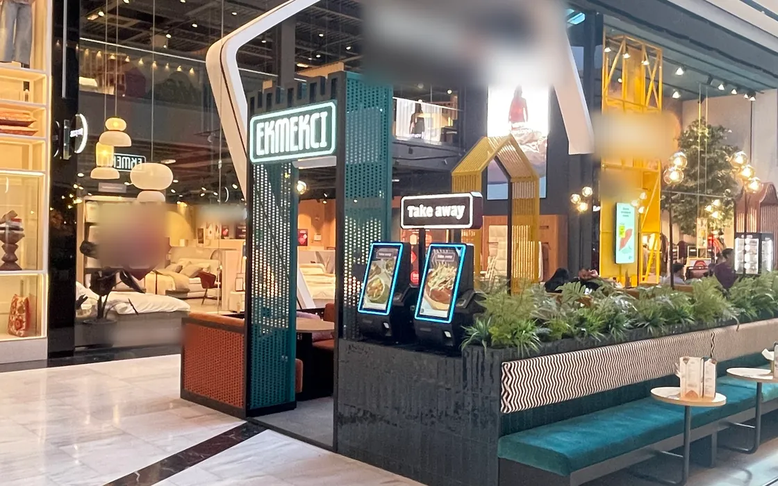 De kiosk van Ekmekci van dichtbij: het EKMEKCI-bord op een petrolkleurige zuil, een bord Take away, twee bestelzuilen, planten en een groene bank met kleine ronde tafels.